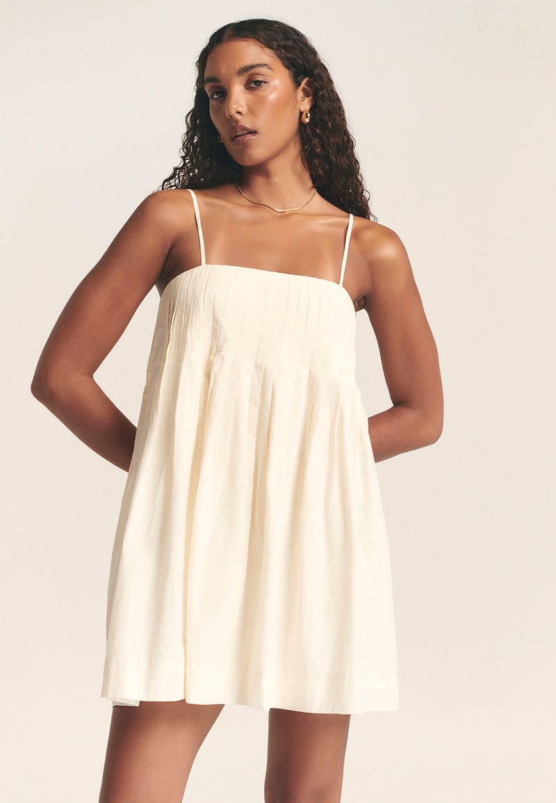 Vestido color crema con finos tirantes tipo spaghetti, que presenta un escote cuadrado y detalles de pliegues verticales. Falda fluida con una textura sutil.