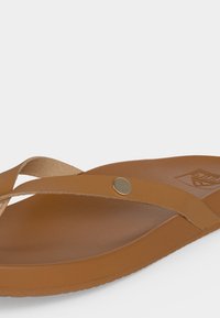 Καφέ δερμάτινο σανδάλι τύπου thong με λεπτό λουράκι, μεταλλικό καρφί και υφασμένη σόλα σε απλό λευκό φόντο.