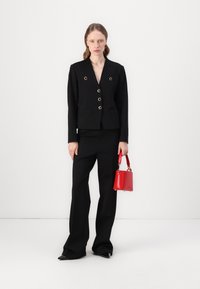 Zwarte blazer met gouden knoopaccenten, gecombineerd met zwarte broeken. Model houdt een kleine rode handtas vast. Eenvoudig, op maat gemaakt ontwerp.