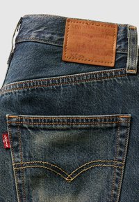 Blugi din denim cu o etichetă din piele bej, cu branding embossat, cusături portocalii și o etichetă roșie pe buzunarul din spate.