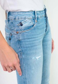 Jean en denim bleu clair avec une coupe slim, des détails légèrement déchirés et plusieurs poches. Le tissu a une texture douce et lisse.