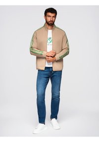 Giacca beige con zip e dettagli verdi sulle spalle, indossata sopra una maglietta bianca e abbinata a jeans blu e sneakers bianche.