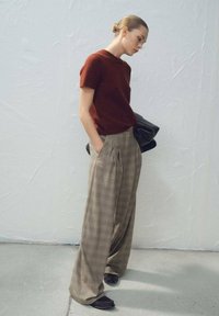 Pull en maille à manches courtes couleur rouille, associé à un pantalon large à carreaux beige et marron. Sac noir porté sur le bras gauche, chaussures foncées.