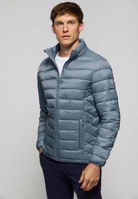 Ljusblå puffjacka med dragkedja, hög krage och horisontell sömnad. Har sidofickor och en logotypapplikation på ärmen.