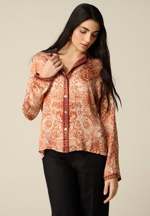 Donna con capelli lunghi e scuri che indossa una camicetta arancione e crema con motivo floreale e bottoni, abbinata a pantaloni neri, posando davanti a uno sfondo semplice.