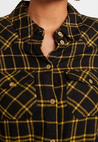 Zwart-geel geruite flanellen overhemd met een button-down kraag, twee borstzakken en bruine knopen. Zachte textuur met een ruitpatroon.