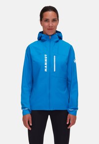 Giacca impermeabile blu con tasca sul petto con zip, cappuccio regolabile e dettagli riflettenti. Presenta un logo prominente sul davanti.