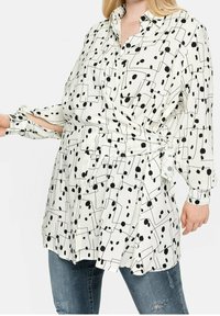 Woman wearing white button-up blouse with black abstract dots and lines, tied at waist, paired with distressed blue jeans.