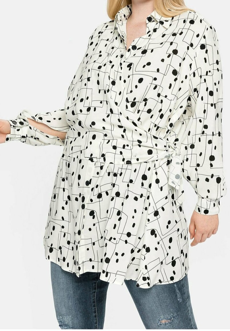 Woman wearing white button-up blouse with black abstract dots and lines, tied at waist, paired with distressed blue jeans.