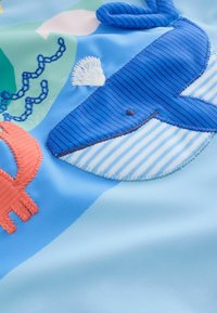 Tessuto blu con balena a fumetti ricamata, pesce arancione e forme verdi. Strisce testurate e aree lisce creano un design giocoso.