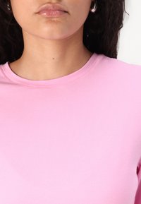 Heled roosa ümar kaelusega T-särk, mis on valmistatud sileda ja veniva kangaga. T-särk on lühikeste varrukatega ja figuuri rõhutava disainiga. Pole silmapaistvaid mustreid ega detaile.