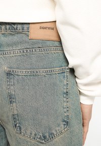 Light blue denim jeans with a faded finish, featuring a brown leather patch labeled "EIGHTYFIVE," and a single back pocket design.