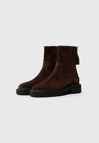 Botine din suede maro, cu o textură netedă, vârf rotunjit și talpă masivă neagră. Au un accent cu ciucure pe spate și design slip-on.