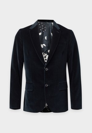Jeune homme portant un blazer en velours noir, une chemise sombre, un pantalon marron à jambes larges et des mocassins bicolores, debout devant un fond blanc.