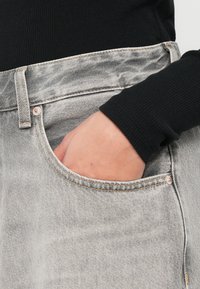 Γκρίζα τζιν με μπροστινές τσέπες, με αντίθετες ραφές και λεπτομέρεια από χαλκό στο κουμπί. Φαίνεται ένα μαύρο ριμπ μπλουζάκι με μακριά μανίκια.