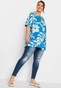 Top floreale blu con maniche oversize abbinato a jeans blu strappati. Modella con sandali bianchi con cinturini. Sfondo semplice.