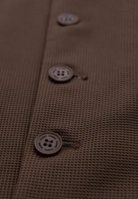 Brauner, strukturierter Stoff mit einem Mesh-Muster, ausgestattet mit drei dunklen Knöpfen, von denen jeder vier Löcher hat, wobei ein Knopf teilweise geöffnet ist.