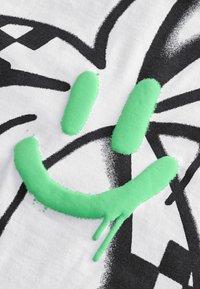 Ein weißer Stoff präsentiert ein strukturiertes, grünes Smiley-Gesicht-Design mit schwarzen grafischen Elementen. Das Smiley hat eine weiche, erhabene Textur und Tropfen.