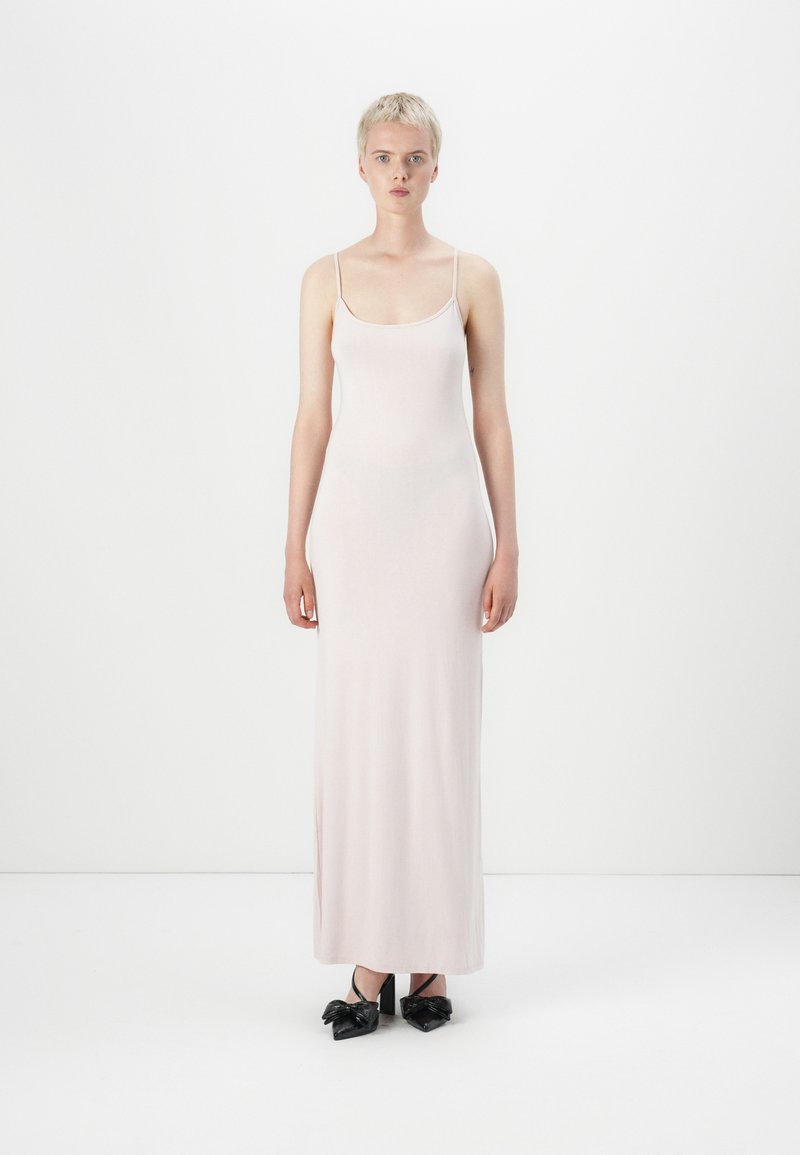 Abito maxi senza maniche di un delicato rosa pallido con spalline sottili, texture liscia e silhouette aderente, abbinato a scarpe con tacco nere con fiocchi.
