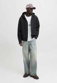 Sort zip-up hoodie med frontlommer, lagd over en hvid t-shirt. Løse lyseblå denimjeans, brune sko med kraftig sål og en lyserød kasket.