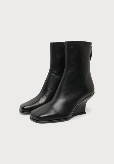 Paire de bottines en cuir noir avec bouts carrés pointus et talons compensés sculptés sur fond blanc.