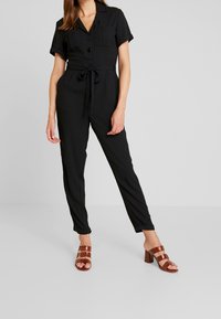 Combinaison noire à manches courtes, avec col, boutons sur le devant et ceinture à nouer. Le mannequin porte des sandales à talons marron avec un design à bretelles.