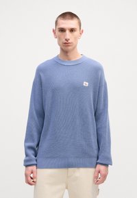 Suéter de malha azul claro com um padrão texturizado, gola redonda e um pequeno patch de logotipo branco no lado esquerdo do peito. Corte solto.