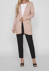 Kvinna iklädd en ljusrosa öppenvävd cardigan, vit topp, svarta smala byxor och svarta sandaletter med öppen tå, stående mot en enfärgad bakgrund.