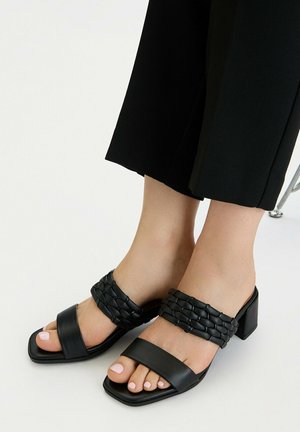 Pies con sandalias negras de tacón ancho con una correa trenzada y una correa frontal lisa, combinadas con pantalones negros recortados.
