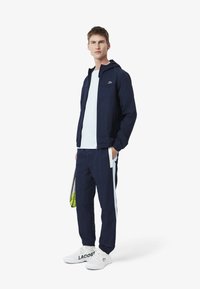 Marineblauwe ritsjack met capuchon en zakken, gecombineerd met bijpassende broek met witte zijkanten. Witte sneakers met logo-detail.