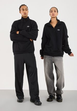 To personer med flettede hår har på seg svarte half-zip hettegensere, løse bukser og svarte sko, og står foran en enkel hvit bakgrunn.