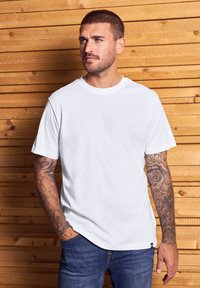 Bílý bavlněný tričko s kulatým výstřihem, krátkými rukávy, volným střihem a bez viditelných vzorů. Nosené s modrými džínsy. Pozadí s dřevěným obložením.