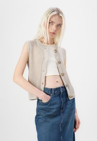 Beige strikket vest med rund halsutringning, ermeløs design og seks treknapper. Bæres over en hvit topp og kombinert med et denimskjørt.