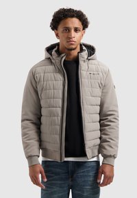 Lichtgrijze gewatteerde jas met een capuchon, voorzien van een ritssluiting aan de voorkant, elastische manchetten en een logo op de borst. Gedragen over een zwarte trui.