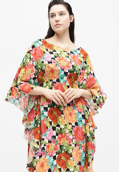 Νέα γυναίκα με σκούρα μαλλιά φοράει πολύχρωμο φόρεμα με λουλούδια και ασπρόμαυρα καρό σε φαρδιά γραμμή, πάνω σε απλό λευκό φόντο.