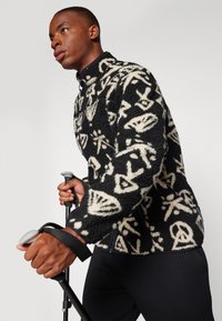Jachetă din fleece de culoare neagră cu modele abstracte crem, guler înalt și închidere cu fermoar. Modelul ține un baston de mers și poartă pantaloni negri.
