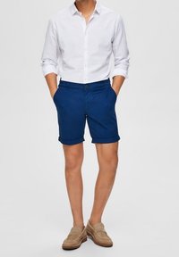 Mäns blå bomullsshorts med uppvikta ärmar, knäppning med knapp, i kombination med en vit långärmad skjorta och beige loafers.