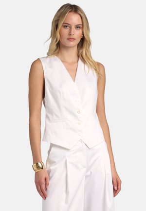 Blondynka w białej kamizelce bez rękawów z guzikami i białych spodniach z plisami, uzupełniona złotą bransoletą mankietową i kolczykami.