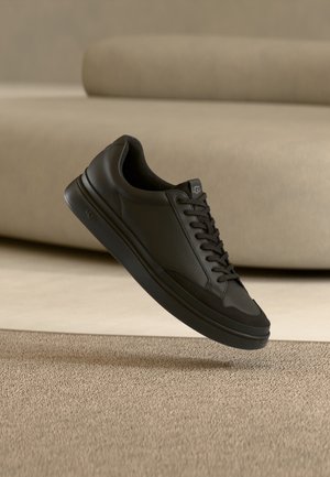 Schwarzer Leder-UGG-Sneaker mit schwarzen Schnürsenkeln, schwebend über beigem Teppich und beigem Sofahintergrund.