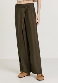 Olivengrüne, weit geschnittene Hosen mit einem Überlappungsdetail an der Vorderseite, kombiniert mit tanfarbenen Mokassins im Fransenstil, die mit Perlenakzenten verziert sind.