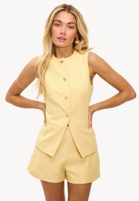 Blazer sans manches jaune sur mesure avec boutons devant, associé à un short assorti ; texture lisse et lignes épurées, avec poches latérales.