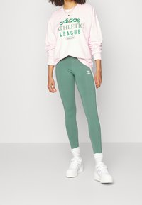 Rosa sweatshirt med grön texten "ATHLETIC LEAGUE", kombinerad med gröna leggings som har vita sidostreck och vita sneakers.