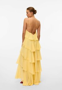 Gul lager-på-lager chiffondress med djup rygg, som har spaghetti-band och flera lager av volanger, vilket ger en flytande silhuett.