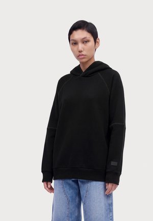 Persona con el cabello negro corto, que lleva una sudadera con capucha negra y vaqueros azul claro, de pie frente a un fondo claro y liso.