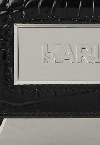 Svart krokodilpräglat läder med en silverfärgad metallplåt som har präglad "KARL"-text och stiliserad liten ansiktsdetalj på K.