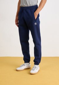 Tumsā zilā krāsā sporta bikses ar elastīgu jostasvietu, sānu kabatām un baltu Adidas logo. Izgatavotas no mīksta auduma ar brīvu piegriezumu un sašaurinātiem piedurknēm.
