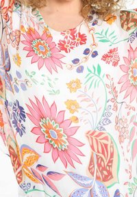 Blusa de estampado floral con fondo blanco, que presenta vibrantes flores rosas, moradas, naranjas y hojas verdes. Tejido ligero y fluido.