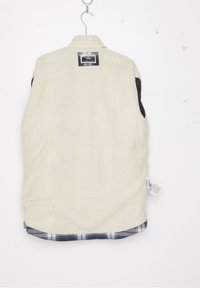 Fleeceväst i krämfärgad med svarta detaljer, med ett rutigt nederkant. Lappen syns på baksidan; mjuk textur och rundad nederkant.