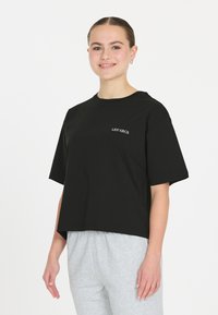 Sort cropped t-shirt med korte ærmer, prydet med hvid tekst "LES ARCS" på fronten. Glat bomuldsstof. Parret med grå sweatpants.