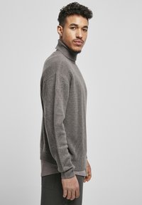 Jersey de cuello alto gris en un tejido suave y texturizado. Presenta un cuello y puños de canalé, y un corte relajado, usado sobre una camisa clara y pantalones oscuros.